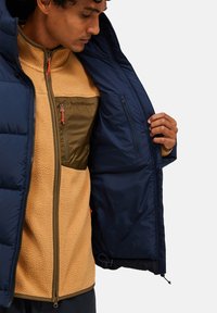 Marineblå isoleret puffjakke over en lys fleece med en lynlåslomme på brystet. Har kontrastsyninger og lyse hårdvare detaljer.