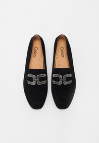 Zwarte suede loafers met een platte zool, voorzien van een decoratieve ketting in zilver en grijze accenten. Het interieur heeft een gevoerd voetbed met perforaties.