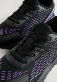 Schwarze Sportschuhe mit lila Akzenten und Mesh-Einsätzen. Merkmale sind eine abgerundete Zehenpartie, eine strukturierte Oberfläche und ein traditionelles Schnürsystem.