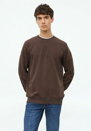 Junger Mann mit kurzen lockigen dunklen Haaren, der einen braunen Pullover mit der Aufschrift "all I need is time and space" und blaue Jeans trägt, vor einem weißen Hintergrund steht.