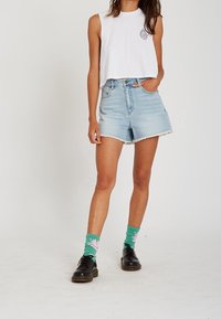 Top blanco corto sin mangas, pantalones cortos de mezclilla de tiro alto en azul claro con orillas deshilachadas, calcetines verdes con patrón floral, zapatos negros.