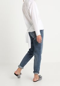 Blusa bianca a maniche lunghe con dettaglio annodato sul retro, abbinata a jeans in denim blu con risvolto e scarpe slip-on grigie a fantasia.