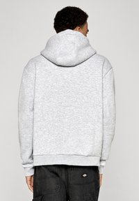 Pelēks vilnas nirto ar lielu, regulējamu kapuci, ribotām piedurknēm un apakšmalu, un bez redzamām grafikām, uzvilkts virs tumšiem kravnieku biksēm.