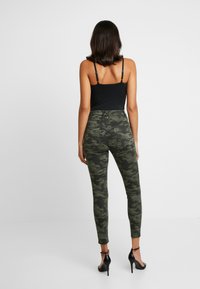 Pantalones de camuflaje de cintura alta en tonos verdes con una silueta ajustada, combinados con una parte superior sin mangas negra y sandalias negras de tacón alto.
