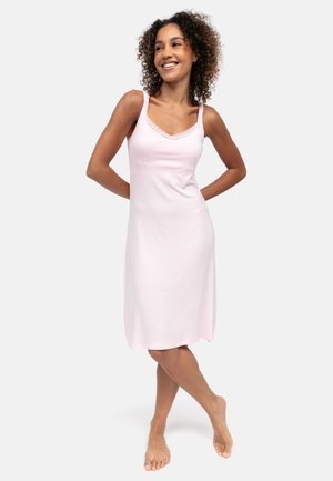 Vrouw met krullend haar, gekleed in een lichtroze mouwloze jurk tot op de knie, staat blootsvoets met haar handen achter haar rug en glimlacht.