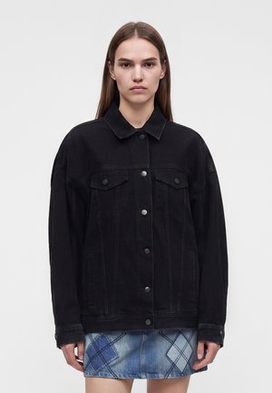 Mulher com cabelo comprido e liso veste um casaco de ganga preto com botões e uma saia de ganga azul em patchwork contra um fundo claro simples.