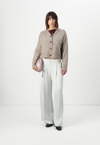 Beslått beige cardigan med store knapper, kombinert med høy midje lysegrønne bukser. Modellen holder en liten lavendelveske. Svarte sko.