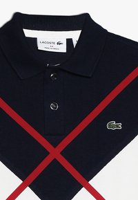 Polo tričko Lacoste s námořnickým modrým pleteným vzorem, které má červený diagonální pruh, dva černé knoflíky a zelené logo krokodýla.