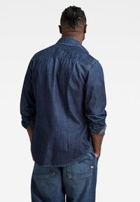 Camisa de mezclilla azul oscuro con mangas largas, canesú en la parte trasera y puños enrollados. Presenta un dobladillo curvado y detalles de costura visibles.
