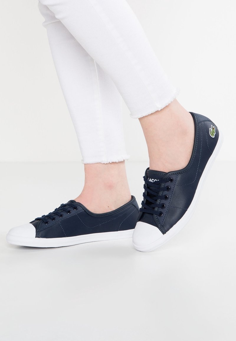 Person som bär marinblå sneakers med vita tåskydd och sålar, i kombination med vita byxor med fransar, står på en ljus yta.