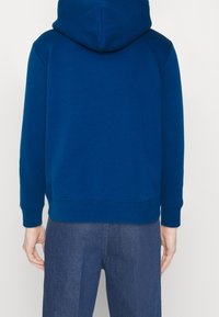 Námořnická modrá mikina s kapucí vyrobená z měkké tkaniny, s klokanní kapsou, žebrovanými manžetami a spodním lemem. Navržena s volným střihem.