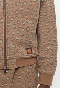 Bruine zip-up hoodie met een stippenpatroon, voorzien van een voorkennis, geribbelde zoom en een Santa Cruz logo patch op links onder.