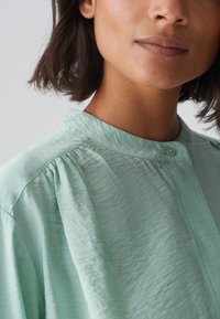 Blusa in tessuto leggero color verde menta, con una texture morbida, design plissettato, colletto alla francese e dettagli con bottoni.