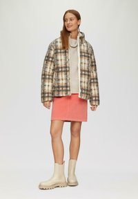 Pufferjack met een beige en geruit patroon, gecombineerd met een lichte trui en een koraalkleurige rok, afgewerkt met beige enkellaarsjes.