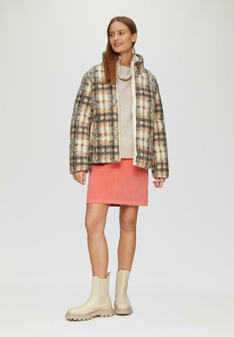Pufferjack met een beige en geruit patroon, gecombineerd met een lichte trui en een koraalkleurige rok, afgewerkt met beige enkellaarsjes.