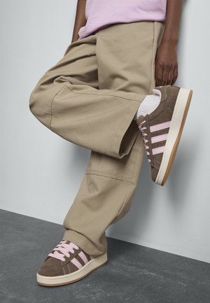 Person som bär beiga vida byxor, vita strumpor och bruna sneakers med rosa ränder, står på ett ben mot en ljus vägg.