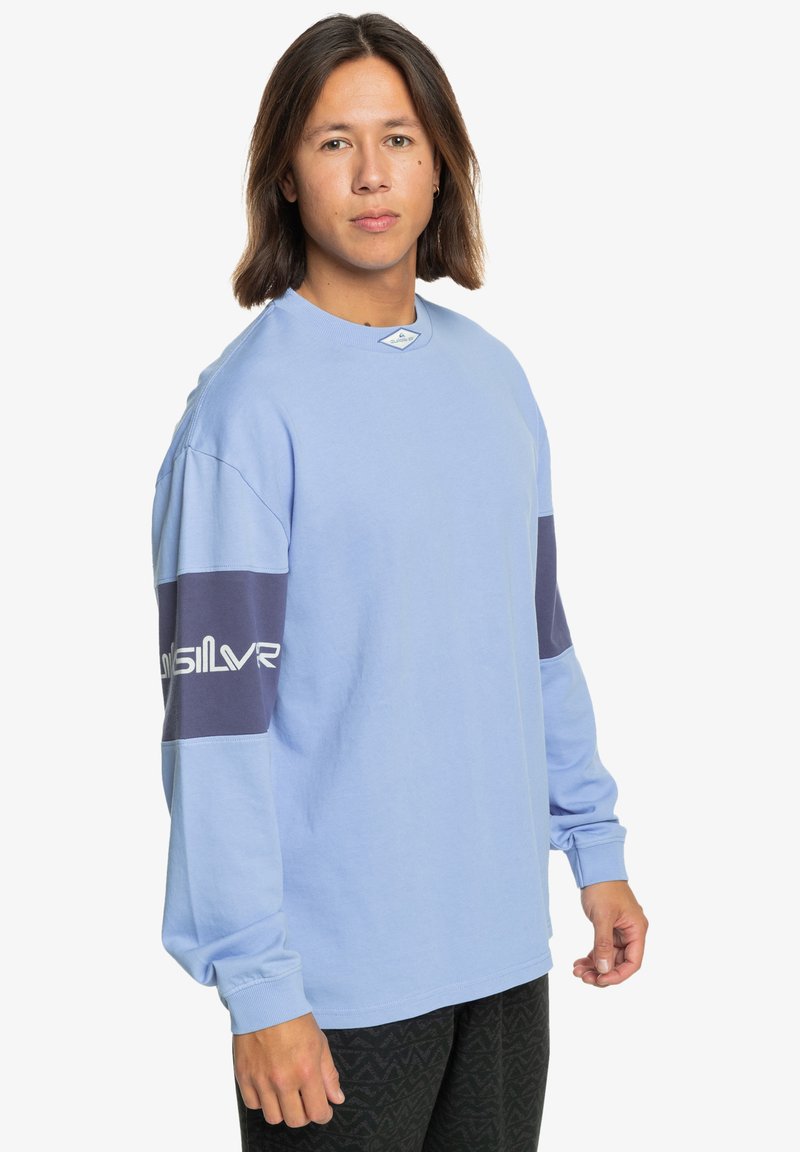 Jasnoniebieska koszula z długim rękawem z granatowymi akcentami na rękawach, z okrągłym dekoltem i detalem logo. Noszona z ciemnymi spodniami w wzory.