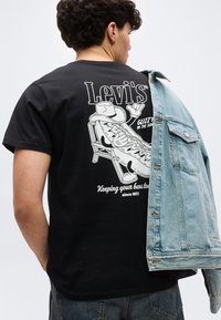 Άτομο που φοράει μαύρο T-shirt Levi's με γραφικό ζεστού σκύλου, ξαπλωμένο σε καρέκλα και ένα τζιν μπουφάν να κρέμεται πάνω σε έναν ώμο.