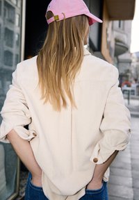 Femme aux longs cheveux blonds portant une casquette rose, une chemise en velours côtelé crème et un jean bleu, debout sur un trottoir en ville, les mains dans les poches.