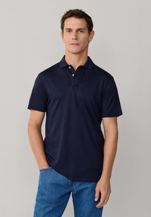 Hombre con camiseta polo azul marino de manga corta con dos botones, vaqueros azules, de pie contra un fondo claro y liso.