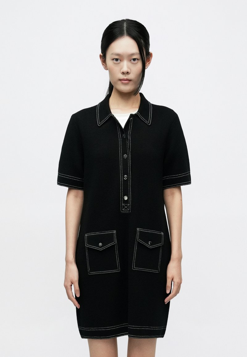 Fekete kötött ruha rövid ujjakkal, kontrasztos fehér varrással, gombos elülső résszel és két első zsebfedővel.