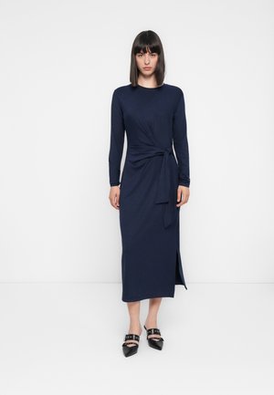 Mujer de pie con el cabello oscuro y liso, vistiendo un vestido midi de manga larga color azul marino con un nudo lateral y zapatos de punta negros.