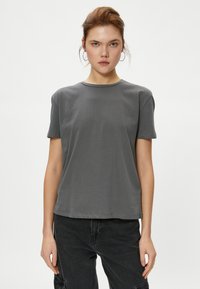 T-shirt en coton gris à manches courtes, col rond et coupe décontractée, présenté sur un mannequin portant un jean foncé devant un fond uni.