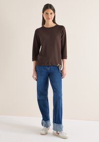 Femme debout portant un haut marron à manches longues avec un détail nœud, un jean bleu retroussé et des baskets blanches, sur un fond clair uni.