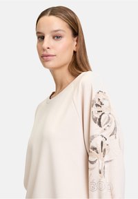Sweatshirt beige clair avec un col rond, présentant des embellissements à motifs sur la manche gauche et le texte "GOD" dans une couleur contrastante.