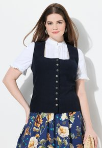 Marine corduroy gilet met vierkante halslijn, versierd met zilveren knopen, gedragen over een witte gerimpelde blouse. Bloemenpatroon rok eronder.