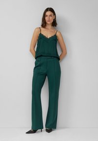 Kobieta stojąca oparta o ścianę, ubrana w ciemnozieloną koszulkę wykończoną koronką, dopasowane spodnie z wysokim stanem oraz czarne szpilki z ostrym noskiem.