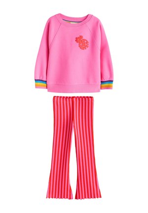 Ung pige smilende, iført en lyserød sweatshirt med regnbuekanter og røde stribede bukser, stående med benene spredt på en beige baggrund.