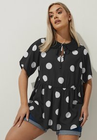 Blusa nera a maniche corte con pois bianchi, colletto e laccio regolabile al collo; vita arricciata con vestibilità ampia.