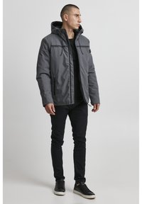 Sivi vodootporni kaput s kapuljačom, prednjim patentnim zatvaračem i dva bočna džepa. Slim fit dizajn s crnim detaljima. Nosi se preko crne majice i hlača.