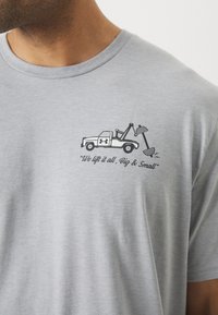 T-shirt en coton gris avec un graphique d'un camion de remorquage soulevant des poids. Le texte indique : "Nous soulevons tout, gros et petit," situé près de la poitrine.