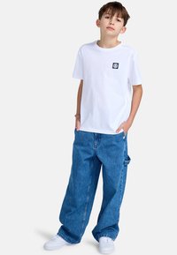 Witte katoenen t-shirt met klein zwart logo op de borst, gecombineerd met losse blauwe denim jeans en witte sneakers.