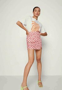 T-shirt branca com gráfico espiral laranja, combinada com uma saia mini xadrez em rosa e branco. Calçado: sandálias de salto alto verdes com tiras.