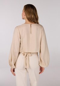 Beige cropped blouse met een textuurafwerking, lange pofmouwen, een sleutelgatopening aan de achterkant en een strikband in de taille. Gecombineerd met lichtgekleurde broeken.