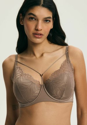 Kobieta o długich ciemnych włosach, ubrana w taupe koronkowy stanik z fiszbinami i ozdobnymi paskami na jednolitym jasnym tle.