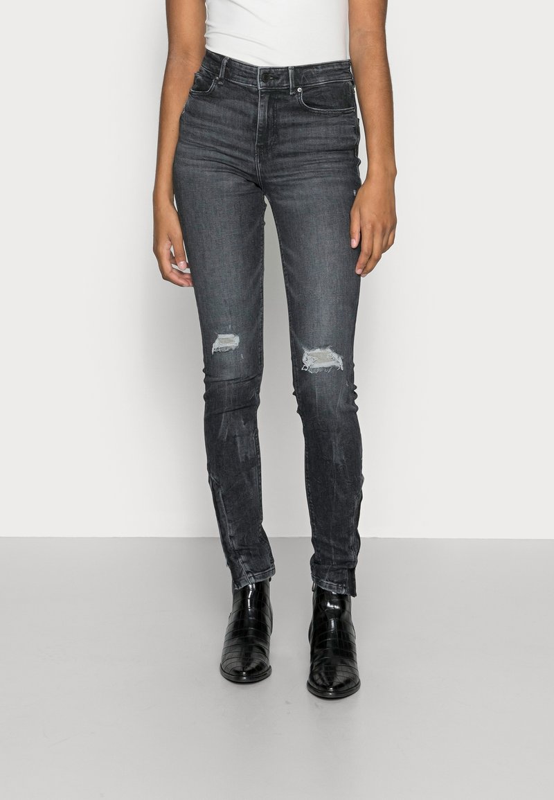 Femme portant un jean skinny taille haute gris foncé avec des patchs usés aux genoux et des bottines noires, debout devant un fond blanc.