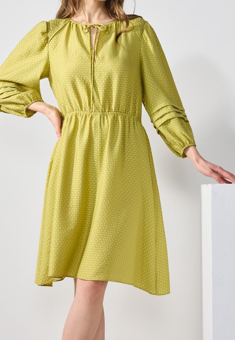 Robe verte texturée avec un col rond, un détail noué et des manches bouffantes. Elle présente une taille cintrée et une jupe fluide avec un ourlet à hauteur des genoux.