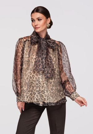 Vrouw met gestileerd donker haar draagt doorschijnende blouse met luipaardprint en grote strik, en gouden opvallende oorbellen, poseert tegen een effen achtergrond.