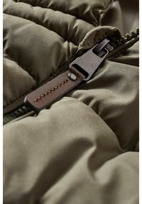 Gros plan sur une fermeture éclair en métal avec une tirette en cuir marron sur une veste matelassée vert olive.