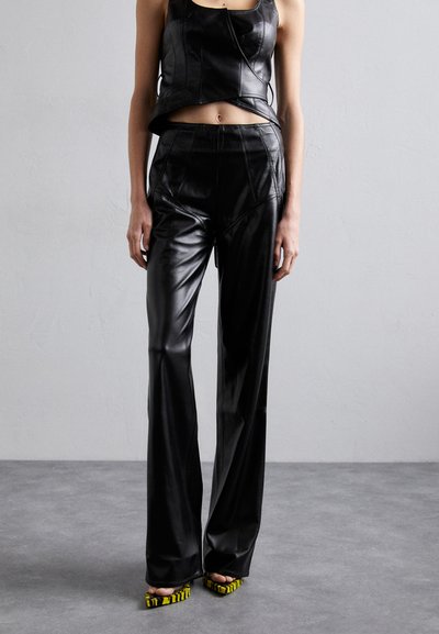 Černý kožený cropped top spárovaný s černými koženými kalhotami s vysokým pasem a rozšířenými nohavicemi. Boty mají žluto-černý pruhovaný design.