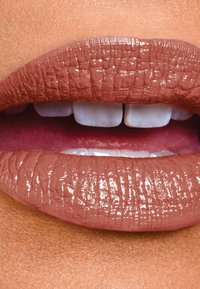 Rouge à lèvres nude mat appliqué sur des lèvres pleines, montrant la texture avec des lignes fines. La finition brillante intensifie la couleur, avec des dents blanches visibles en arrière-plan.