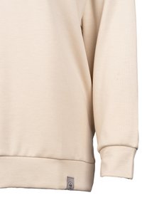 Beżowa bluza z miękką strukturą, o luźnym kroju, z prążkowanymi mankietami i małą metką z logo przyszytą na dolnym dolnym końcu.