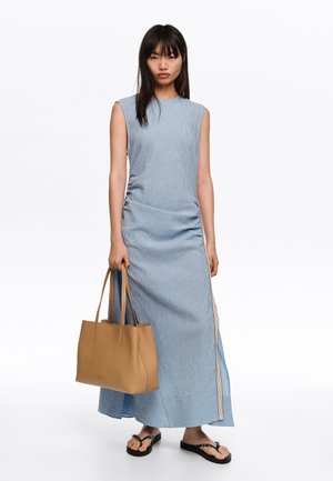 Naine, kes kannab varrukateta sinist triibulist maksikleit, hoiab käes tan-värvi nahast käekotti ja jalas tal mustad varbavahedega plätud, seisab valge tausta ees.