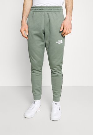 Mężczyzna w oliwkowozielonych dresach The North Face i białych butach Adidas, stojący na tle gładkiego, jasnego tła.