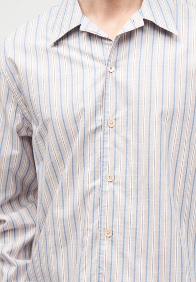 Άνδρας που φοράει πουκάμισο σε μπεζ και ανοιχτό μπλε με κάθετες ρίγες, με τέσσερα ορατά κουμπιά και μυτερό γιακά.