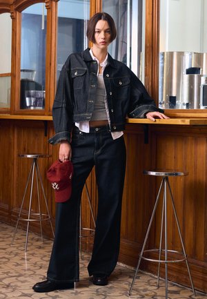 Tânără în jachetă și pantaloni negri din denim, sprijinită de un tejghea de lemn, ținând o șapcă roșie, stând între două scaune metalice în interior.